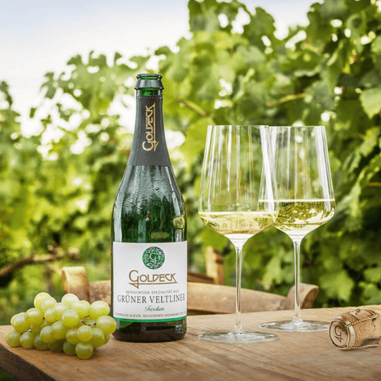 Goldeck Grüner Veltliner steht gemeinsam mit zwei gefüllten Gläsern auf einem Holztisch. Im Hintergrund sind Weinreben zu sehen.